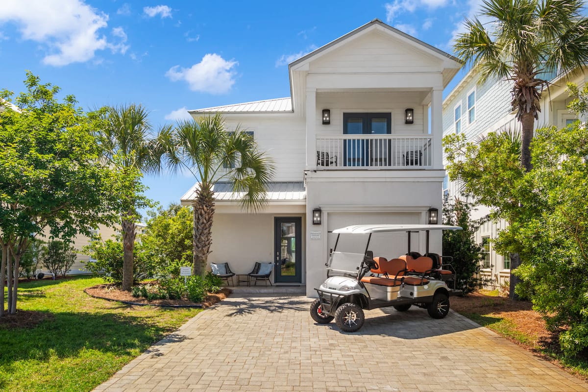 Modern beach house with palm trees and a golf cart parked in the driveway under a bright blue sky.