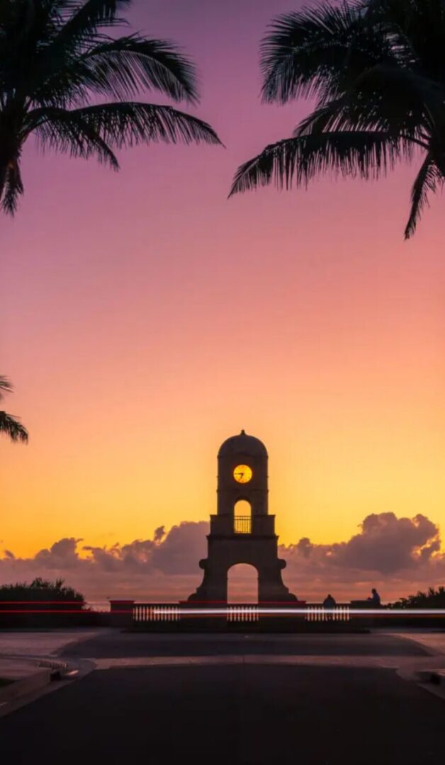 Silhouetted clock tower against a vibrant sunset, framed by palm trees, highlighting the charm of the neighborhood.