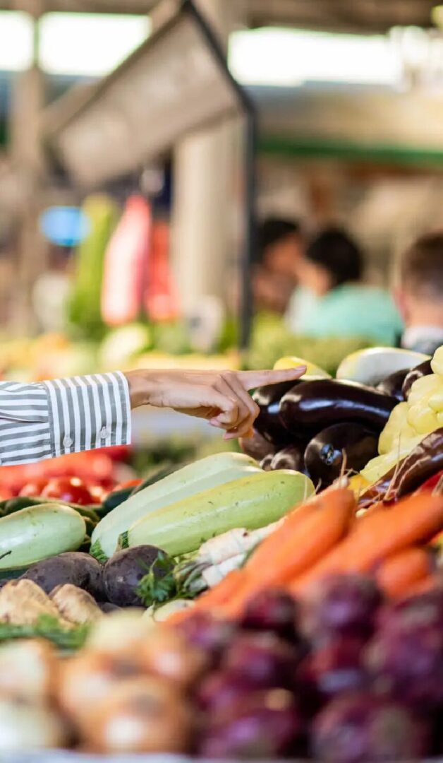 A hand points at a colorful display of fresh vegetables in a bustling market setting.