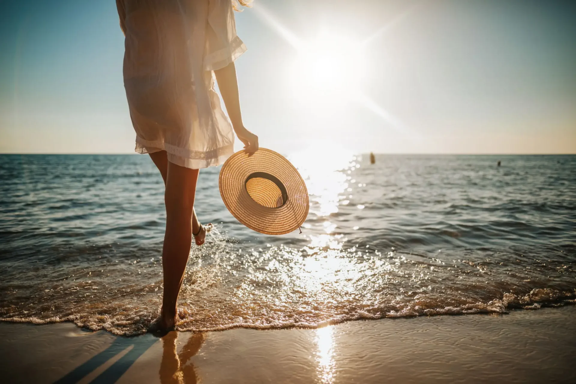 Person walking along sunny beach holding straw hat